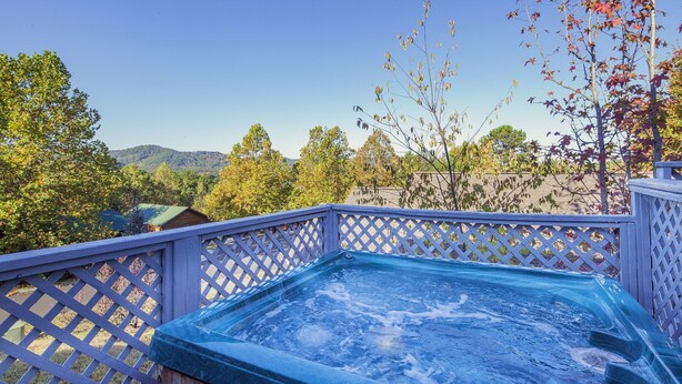 A hot tub on a wooden deck with a white lattice railing, surrounded by trees and a mountainous backdrop. - Our Tennessee Mountain Home - Great Location!  Close To All The Action! (Pigeon Forge)