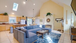 A spacious living room with a large gray sectional sofa, a coffee table, and a blue rug. The kitchen is visible in the background with wooden cabinets and a refrigerator. - #119 Sunshine Shores (Pacific City)