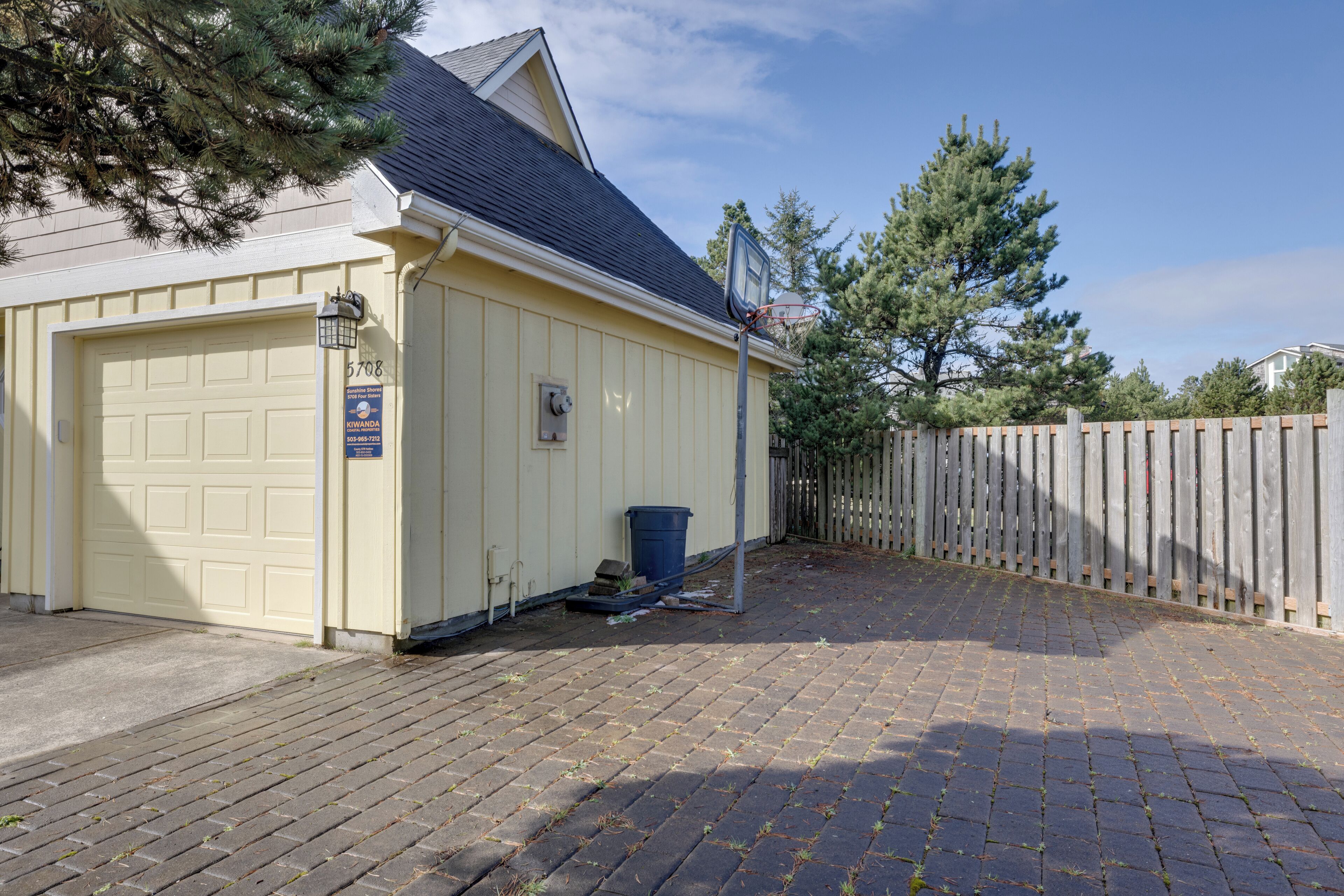 A garage with a blue trash bin, a wooden fence, and a paved driveway.