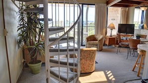 En spiraltrappe fører op til en balkon med havudsigt, et fjernsyn og et spiseområde med fletmøbler.