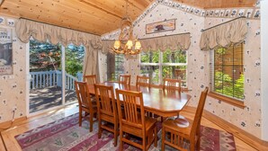 Un espace repas comprenant une table et des chaises en bois, de grandes fenêtres équipées de stores et un plafond avec des poutres apparentes.