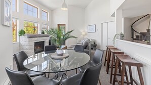 Una habitación amplia con una mesa redonda de vidrio rodeada de sillas, una chimenea y una escalera que sube a un nivel superior.