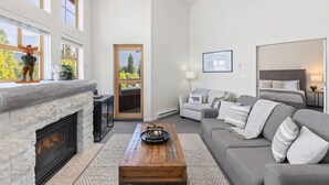 Un salón amplio con chimenea, un conjunto de sofás, una mesa de centro y una puerta que da a un balcón con vistas a árboles.
