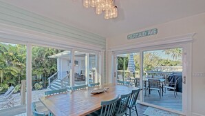 Un comedor con mesa y sillas de madera, vista a una terraza con mobiliario exterior y un cartel que dice "MERMAID AVE".