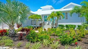 Una casa blanca con un porche cubierto, rodeada de diversas plantas tropicales y un cielo azul despejado.
