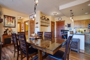 A dining area with a glass table surrounded by chairs, a kitchen area with wooden cabinets, and a clock on the wall.
