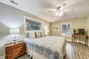 Quarto com cama, penteadeira, ventoinha no teto, suporte para TV e duas janelas.