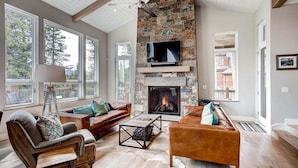 A spacious living room with a stone fireplace, a television mounted on the wall, and large windows offering a view of the outdoors. - Sunrise Ski Haus: Peak 8 Breckenridge Home with Hot Tub and Views, Near Slopes (Breckenridge)