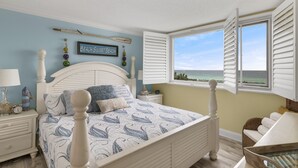 Un dormitorio con cama, mesita de noche, lámpara, ventana con contraventanas y vista a la playa.