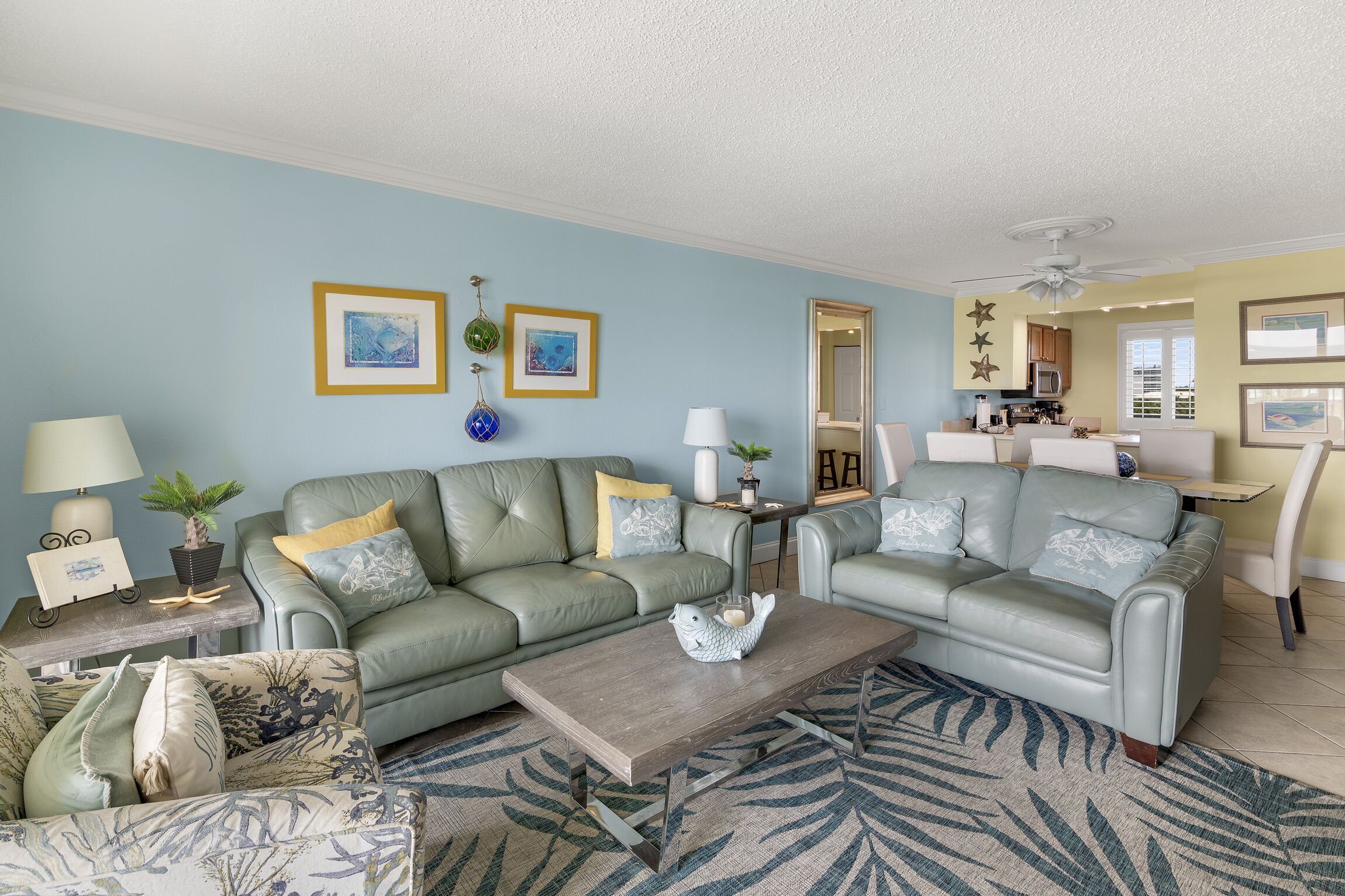 A living room with two green sofas, a coffee table, and a patterned rug. There are framed pictures on the wall and a ceiling fan.