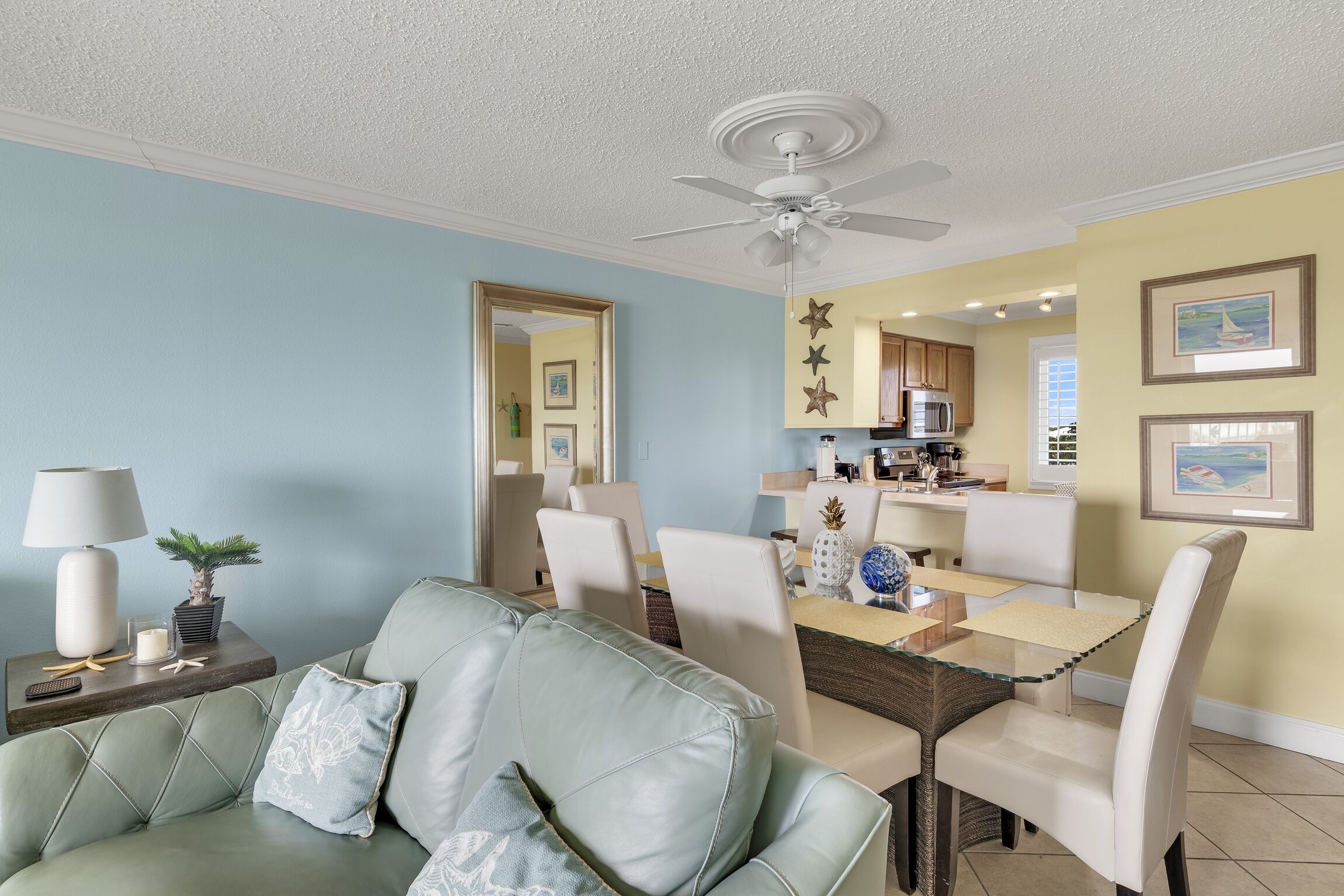 Un salon avec un canapé vert, une table à manger avec des chaises, un ventilateur de plafond et, au fond, un coin cuisine.