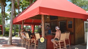 Un espace bar extérieur avec des chaises en bois et un parasol rouge, un panneau noir annonçant les boissons et les plats proposés, et une structure en bois.