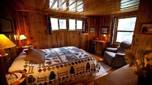 A wooden cabin room with a bed, a chair, a small table, and a window. - Montgomery's Cozy Forest Retreat (Greer)
