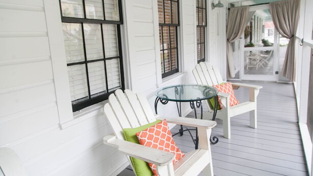 A covered porch with white wooden railings, two white Adirondack chairs with patterned cushions, and a glass table. - Screened Inn c1925! Quiet North End Duplex! Screened porch. Beach Access! (Tybee Island)