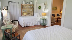 A bedroom with a bed, a chair, a small table, a bookshelf, and a door leading to another room. - Screened Inn c1925! Quiet North End Duplex! Screened porch. Beach Access! (Tybee Island)