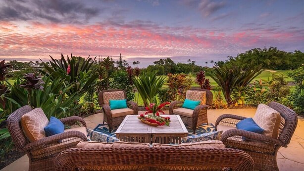 An outdoor seating area with wicker furniture and a central table, surrounded by lush greenery and a scenic view. - Sunlit Luxe Home ~ Ocean Vista + Sunsets + Full AC (Princeville)