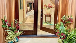 A wooden door leading to a hallway with a staircase, a vase with flowers, and potted plants. - Sunlit Luxe Home ~ Ocean Vista + Sunsets + Full AC (Princeville)