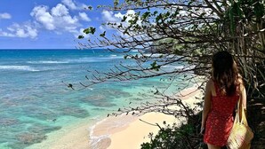 A woman in a red dress standing on a beach with clear turquoise water and a sandy shore. - Sunlit Luxe Home ~ Ocean Vista + Sunsets + Full AC (Princeville)