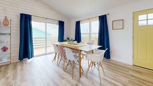 A dining area with a wooden table, white chairs, and blue curtains. - Ocean View from Back Porch, Boardwalk, Outdoor Shower (Port Aransas)