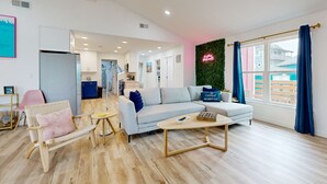 A modern living room with a sofa, a coffee table, and a wicker chair. There is a window with curtains and a neon sign on the wall. - Ocean View from Back Porch, Boardwalk, Outdoor Shower (Port Aransas)