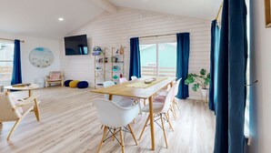A spacious room with a wooden dining table, white chairs, and a large window with curtains. - Ocean View from Back Porch, Boardwalk, Outdoor Shower (Port Aransas)