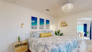 A bedroom with a bed, a bedside table, wall art, a chandelier, and a view of another room. - Ocean View from Back Porch, Boardwalk, Outdoor Shower (Port Aransas)