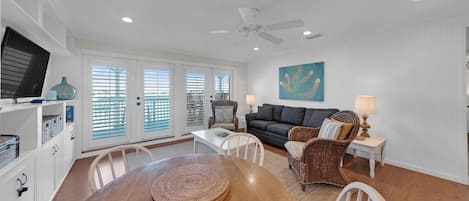 A spacious living room with a wooden dining table, white chairs, a dark grey sofa, and a wicker chair. There is a television on a white cabinet, a painting on the wall, and a ceiling fan.