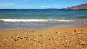 Una playa con agua azul cristalina, piedras y una montaña a lo lejos.