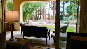Ein Zimmer mit einer Couch, einer Lampe und Blick auf einen angelegten Garten mit Palmen.