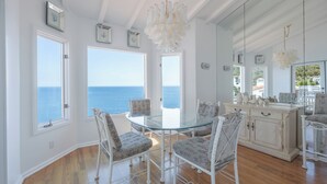 Un comedor con mesa de vidrio y sillas blancas, ventanas amplias con vista al mar y un armario blanco con objetos decorativos.