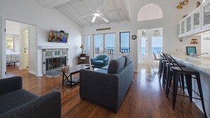Amplia sala de estar con chimenea, un sofá, una mesa de centro y un televisor montado en la pared. Cuenta con una zona de cocina con barra y taburetes, y grandes ventanales con vista al océano.