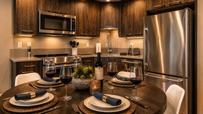 A modern kitchen with dark wood cabinets, stainless steel appliances, and a dining table set for six with wine glasses and a bottle.
