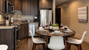 Uma cozinha moderna com armários de madeira escura, eletrodomésticos de aço inoxidável e uma sala de jantar com uma mesa redonda para quatro pessoas.