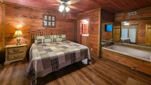 A wooden cabin bedroom with a bed, a nightstand, a lamp, a ceiling fan, a large mirror, and a bathtub. - ER 23 – Smoky Memories  Great Location – Close to town! (Pigeon Forge)