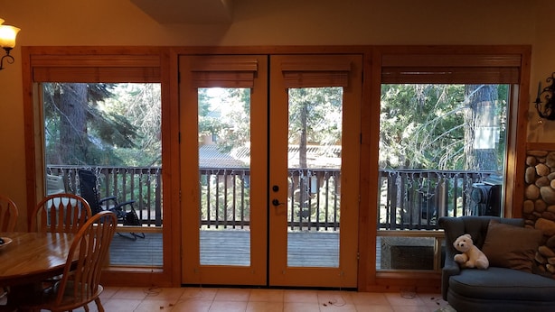 A room with a wooden floor, a dining table with chairs, a sofa with a teddy bear, and a view of a wooden deck and trees through large glass doors.