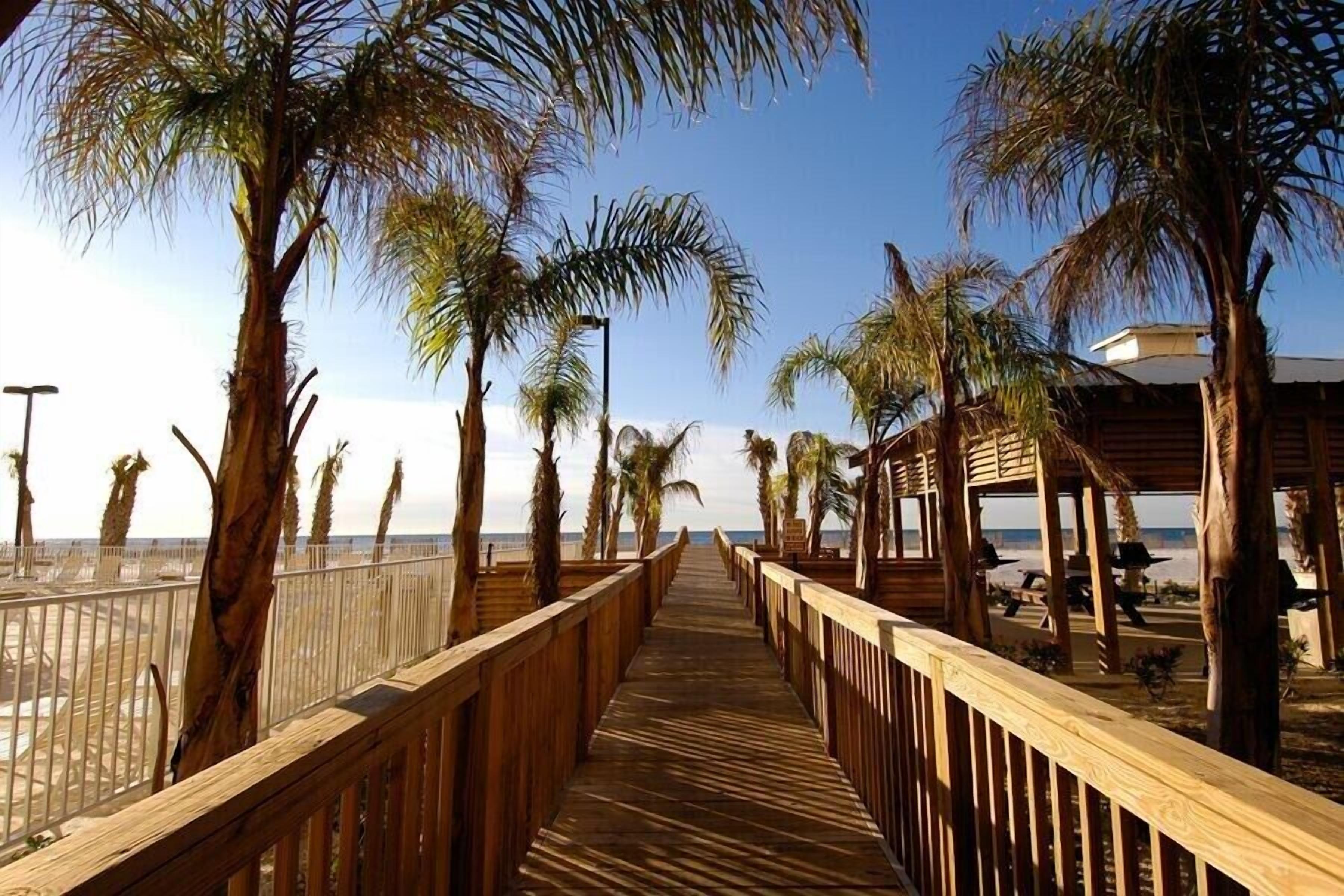 A wooden boardwalk leading to a beach area with palm trees and a covered seating area.
