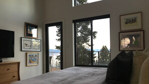Ein Schlafzimmer mit einem großen Bett, einer Kommode, einem Fernseher und einem Fenster mit Blick auf Bäume und ein Gewässer.
