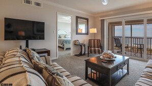 A living room with a television, a sofa, a coffee table, and a chair. There is a balcony with a view of the beach. - Sand Caper Beachfront Condo With Spectacular 7th Floor View of the Gulf! (Ft. Myers Beach)