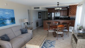 A living room with a sofa, coffee table, and patterned rug. There is a kitchen area with cabinets and a refrigerator. - Oceanfront corner Condo Stunning Views -heated pool Satellite Beach -Cocoa Beach (Satellite Beach)
