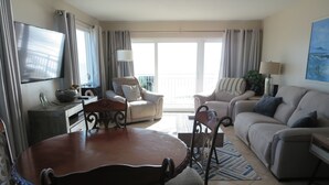 A spacious living room with a dining table, chairs, sofas, and a TV. - Oceanfront corner Condo Stunning Views -heated pool Satellite Beach -Cocoa Beach (Satellite Beach)
