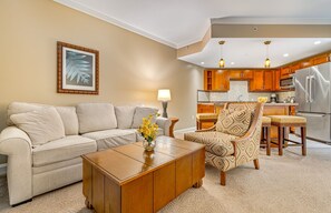 A living room with a sofa, a patterned armchair, a coffee table, and a kitchen area with wooden cabinets and a refrigerator.