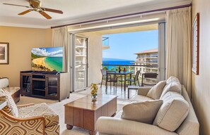 A hotel room with a balcony, a television, a sofa, and a dining area with a view of the ocean.