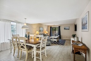 Een eethoek met een houten tafel en stoelen, een bank en een salontafel. Er is een televisie op een standaard en een ingelijste foto aan de muur.