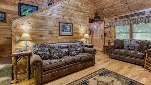 A cozy log cabin interior with a central brown sofa, two side tables with lamps, a wooden staircase, and a large window with curtains.
