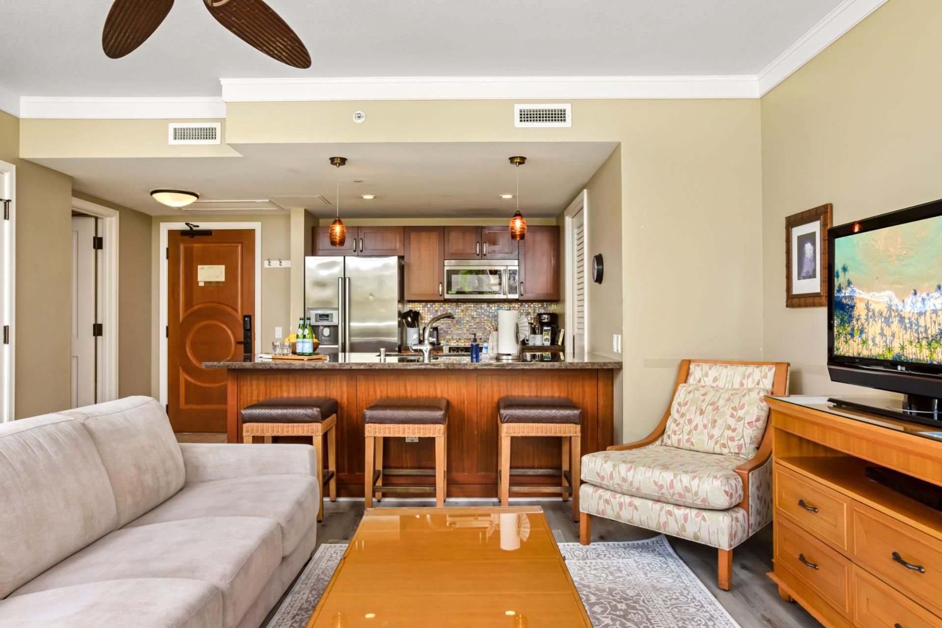 A compact living space with a kitchenette, a sofa, a bar stool, a patterned armchair, and a television.