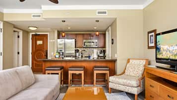 A compact living space with a kitchenette, a sofa, a bar stool, a patterned armchair, and a television.