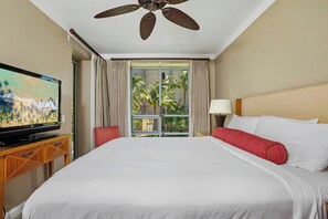 A hotel room with a bed, a TV, a ceiling fan, a window with curtains, and a view of palm trees.