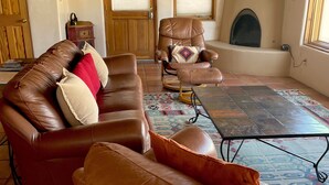 Una sala de estar con un sofá de cuero marrón, un sillón a juego y una alfombra estampada. Se observa una chimenea y una puerta de madera al fondo.