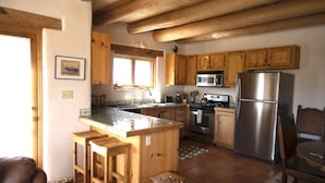 Cocina con armarios de madera, un refrigerador de acero inoxidable, una estufa y un lavabo. Hay dos taburetes de madera y un sofá de cuero marrón.