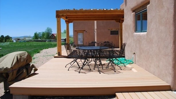 Un patio exterior con terraza de madera, una mesa redonda con sillas y una estructura de pérgola.